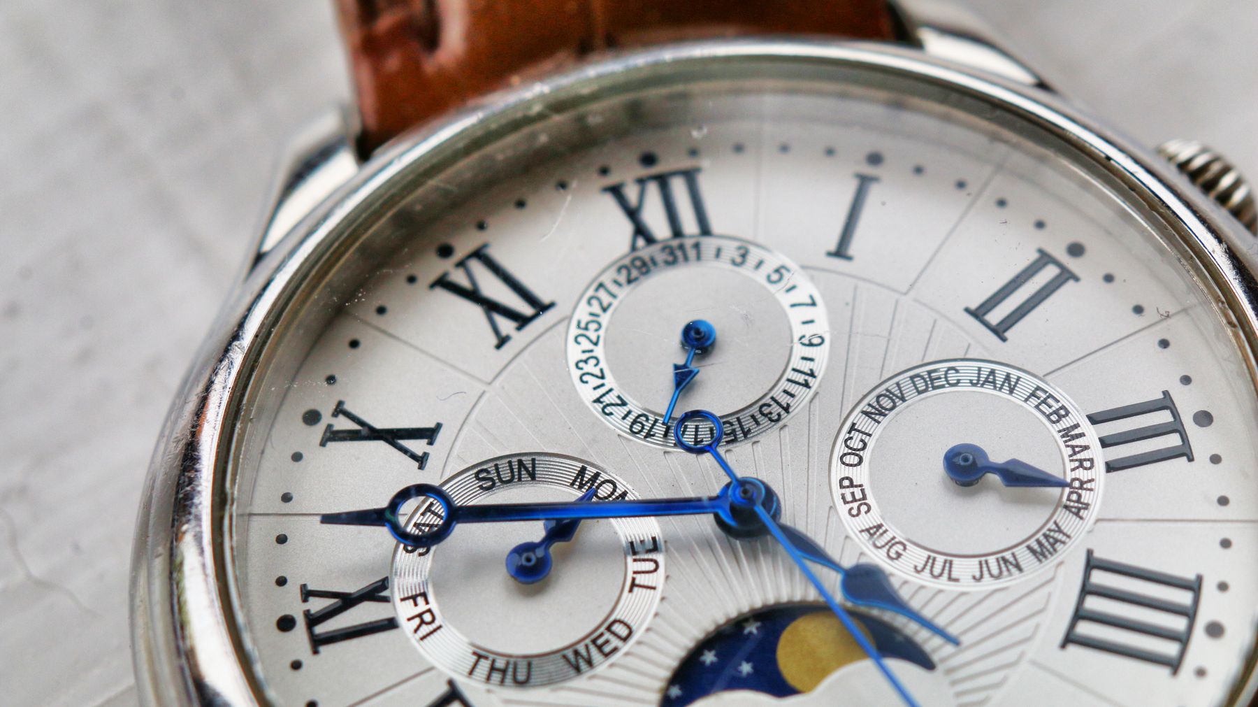 Close up of a white dial with Roman numerals, day and month subdials, a date ring and a moon phase
